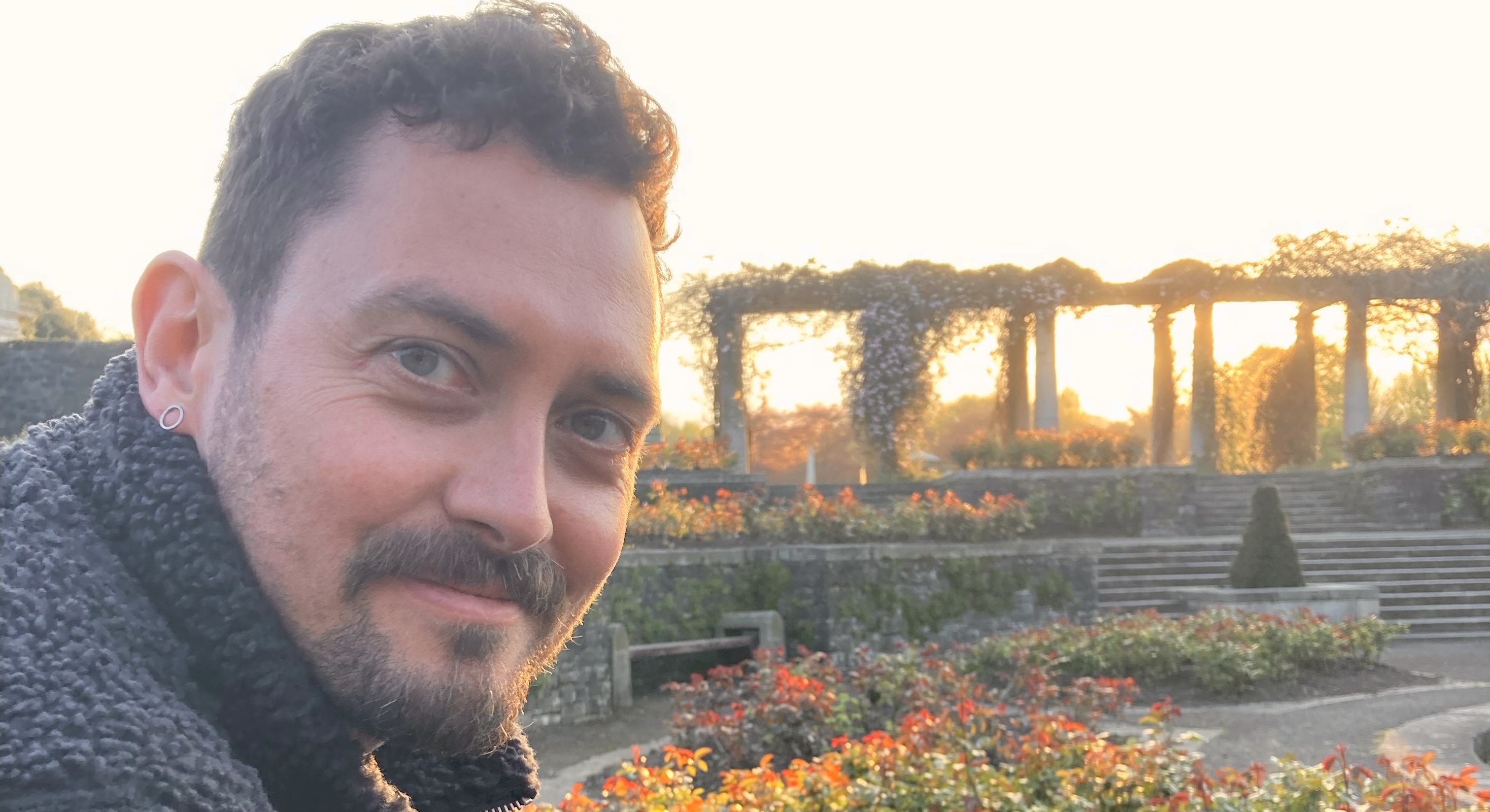 The image shows a man with short curly hair and a beard, smiling while seated in an outdoor garden at
sunset. The background features blooming flowers and a structure with columns entwined with climbing plants.